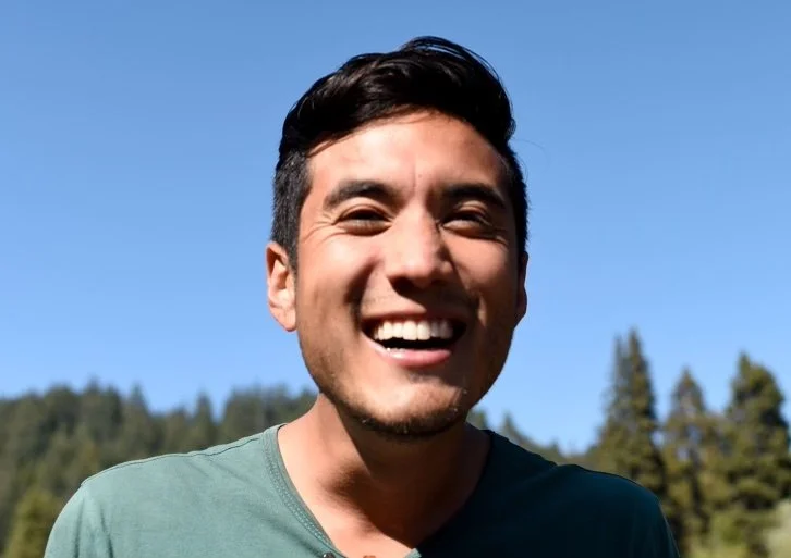A smiling young man outdoors with a blue sky and trees in the background.