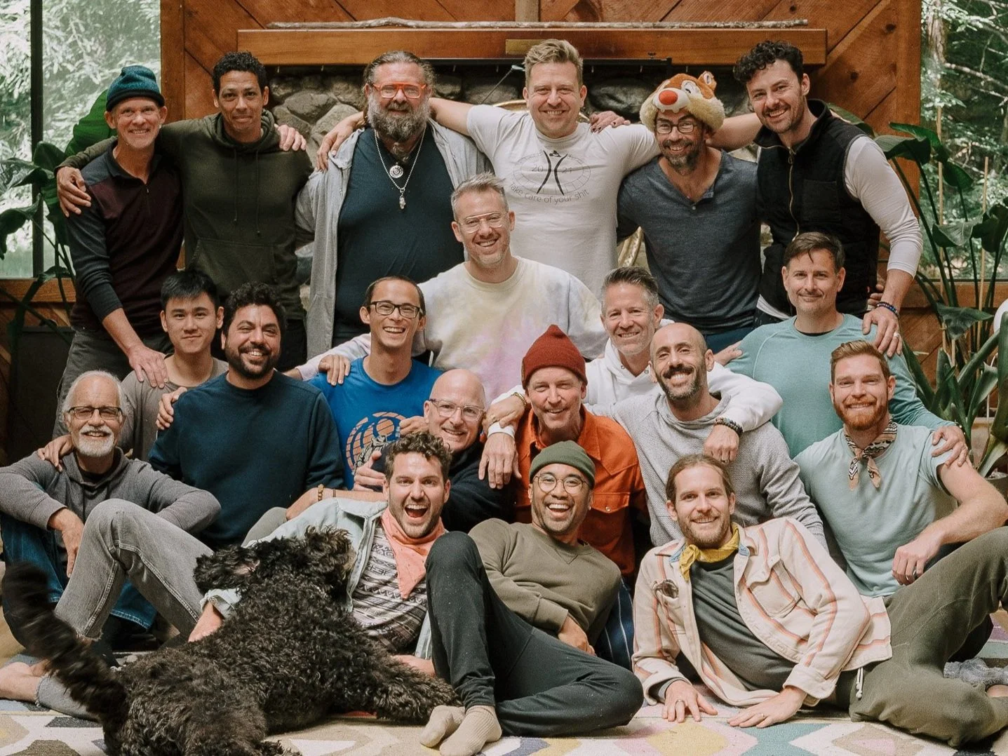 Group of 21 men and one dog posing together indoors, in front of a stone fireplace and surrounded by plants.