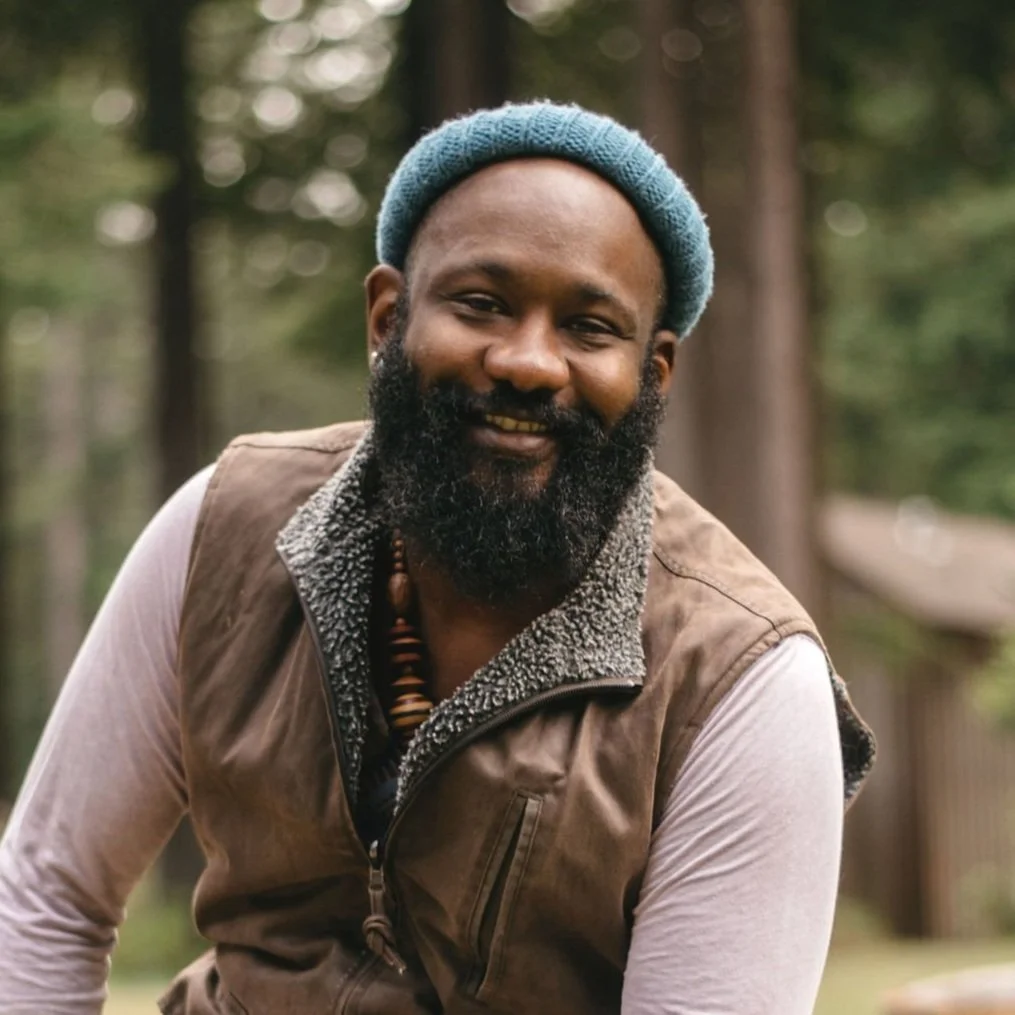 A man outdoors with trees in the background, wearing a blue beanie, a brown vest over a white shirt, and a beaded necklace, smiling at the camera.