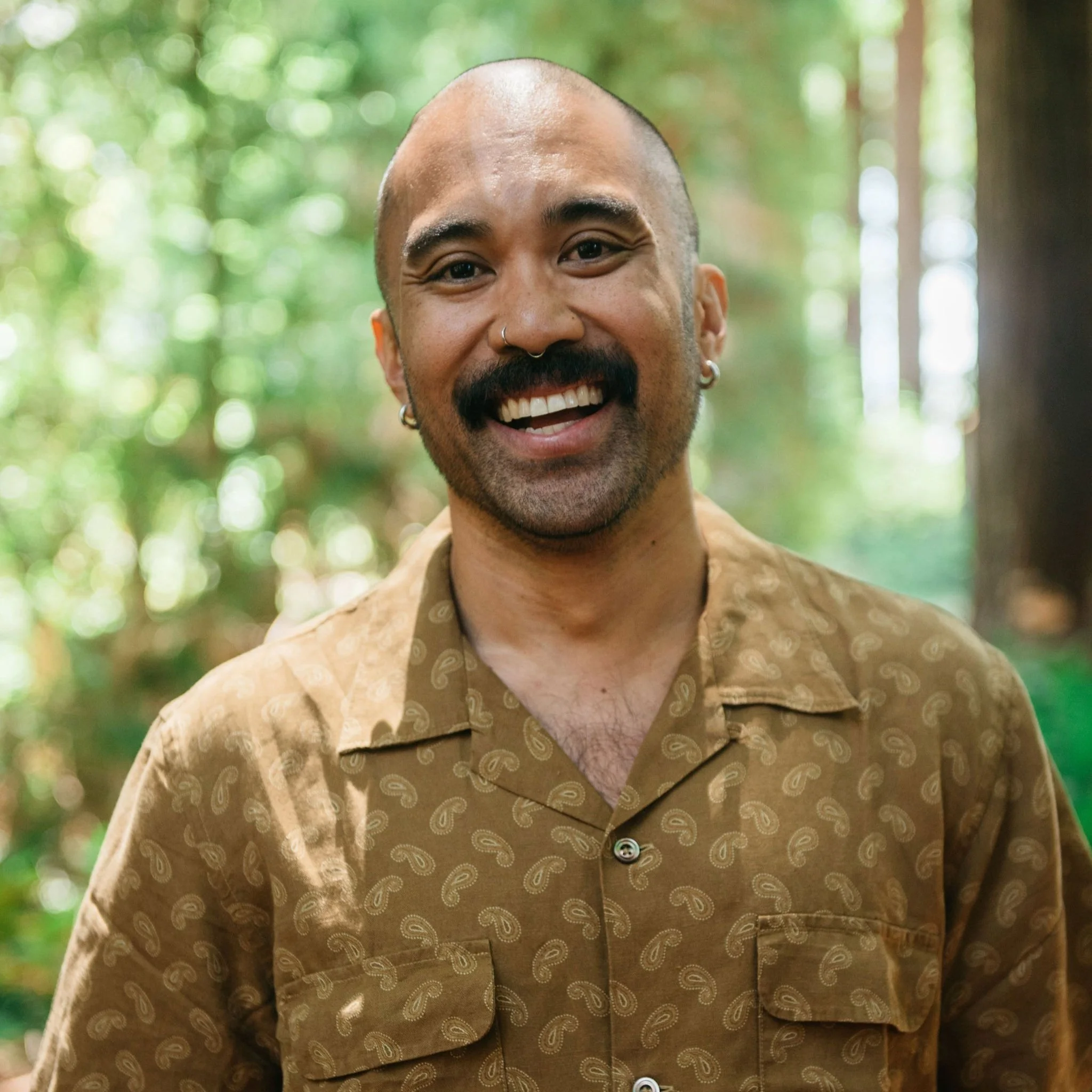 A smiling man with a mustache, beard, and various earrings, standing outdoors in a forest with green trees in the background.