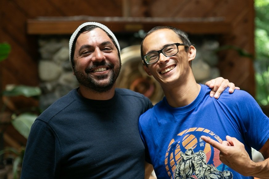 Two men smiling and posing with their arms around each other inside a wooden cabin, with greenery visible through a window in the background.