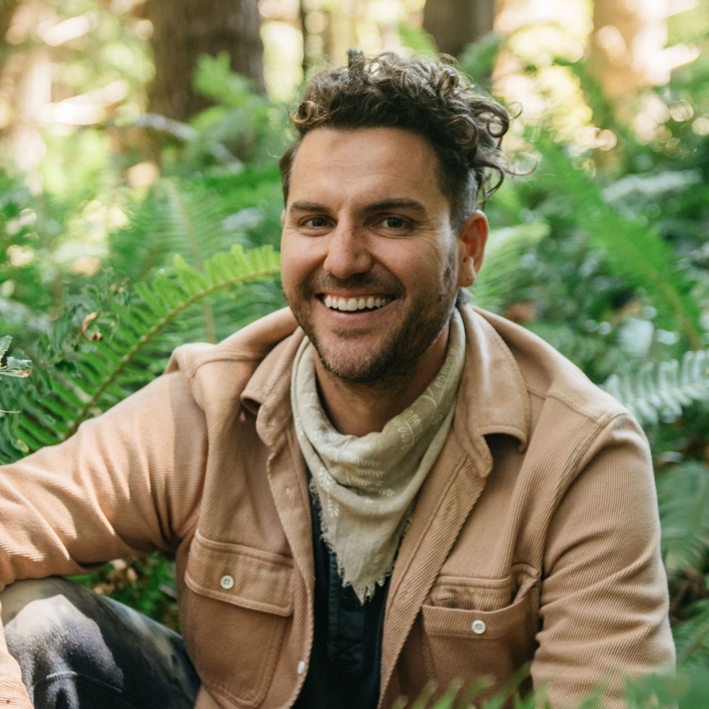 A man smiling outdoors in a lush green forest with ferns, wearing a beige jacket and a bandana around his neck.