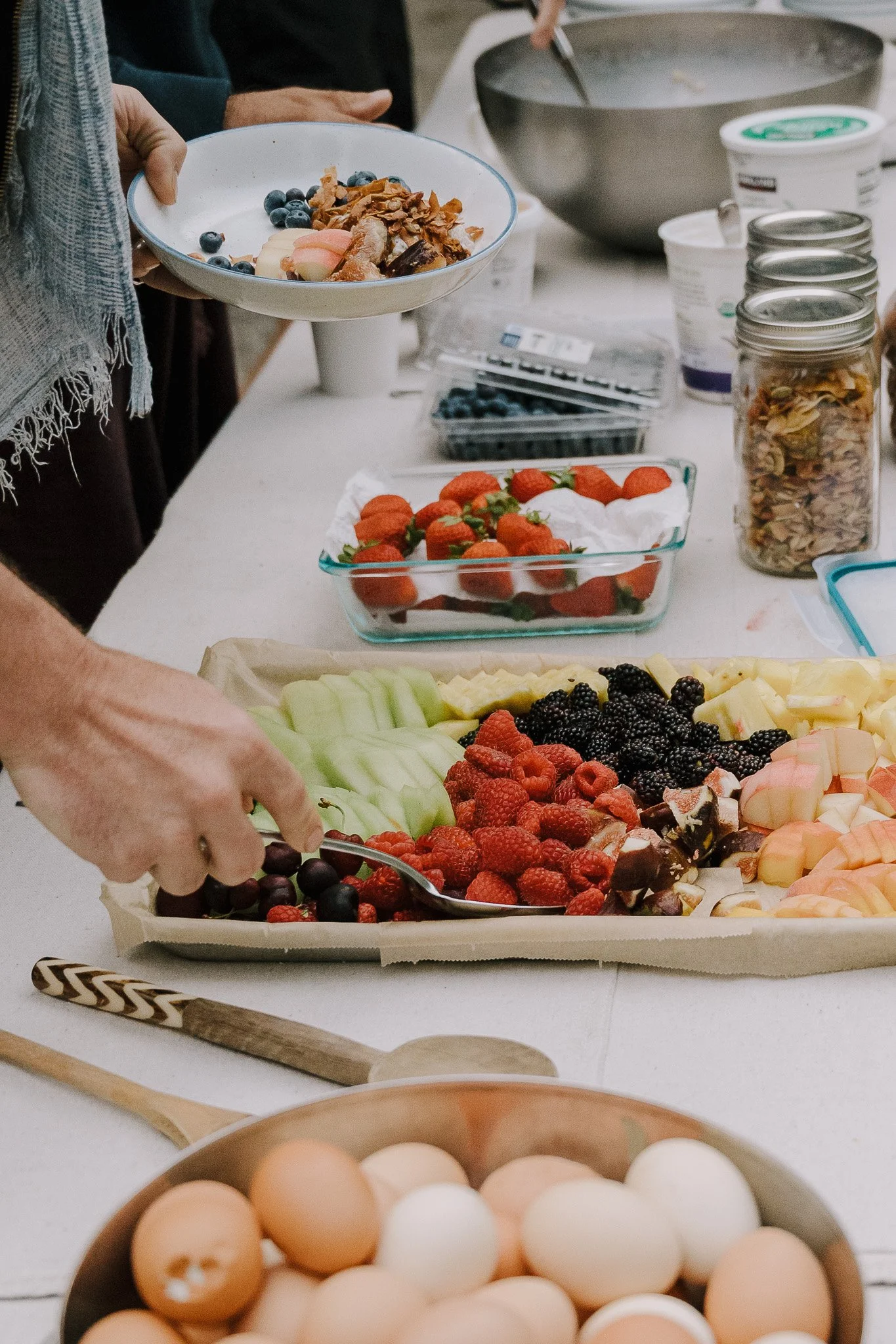 A table with various fresh fruits including strawberries, raspberries, blackberries, honeydew, and other cut fruits, and eggs in a bowl. A person is holding a bowl of fruit salad, and other containers of berries and nuts are on the table.