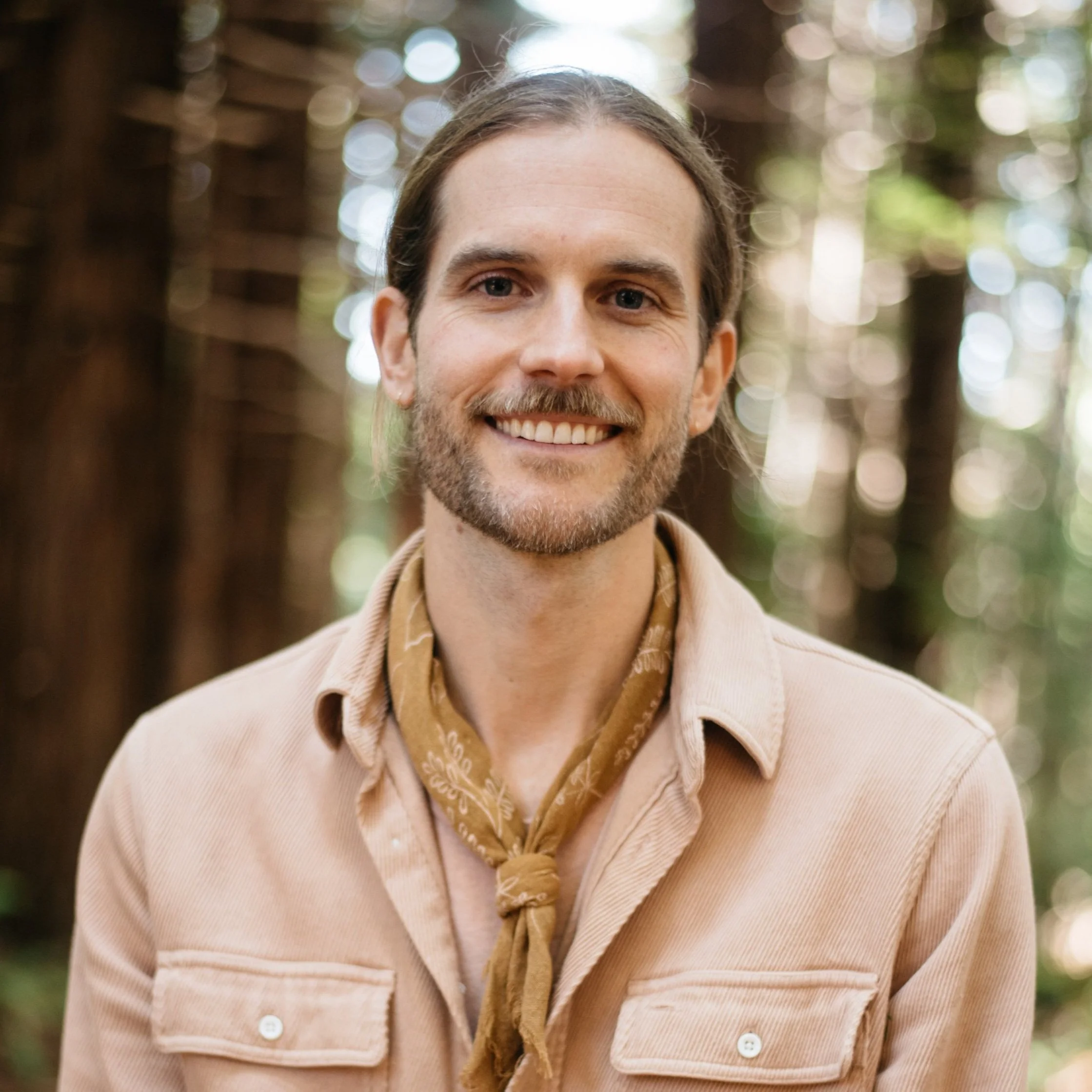 A smiling man with long hair and a beard, wearing a tan shirt and a brown bandana around his neck, standing in a forest with blurred trees in the background.