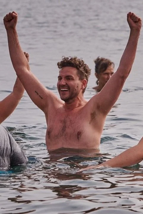 Man celebrating in a body of water with arms raised and a smile, other people visible in the water.