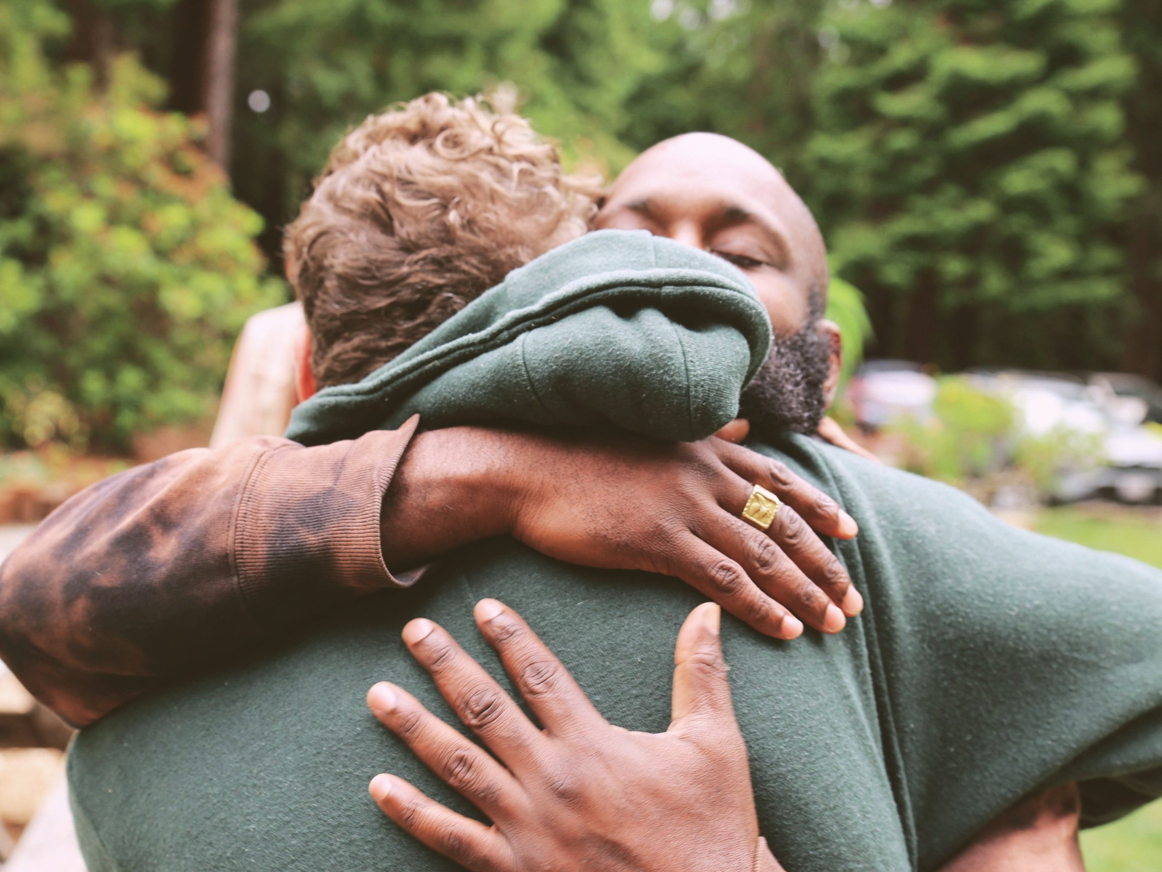 Two men embracing outdoors with greenery in the background, one with light skin and curly hair, the other with darker skin and a beard.