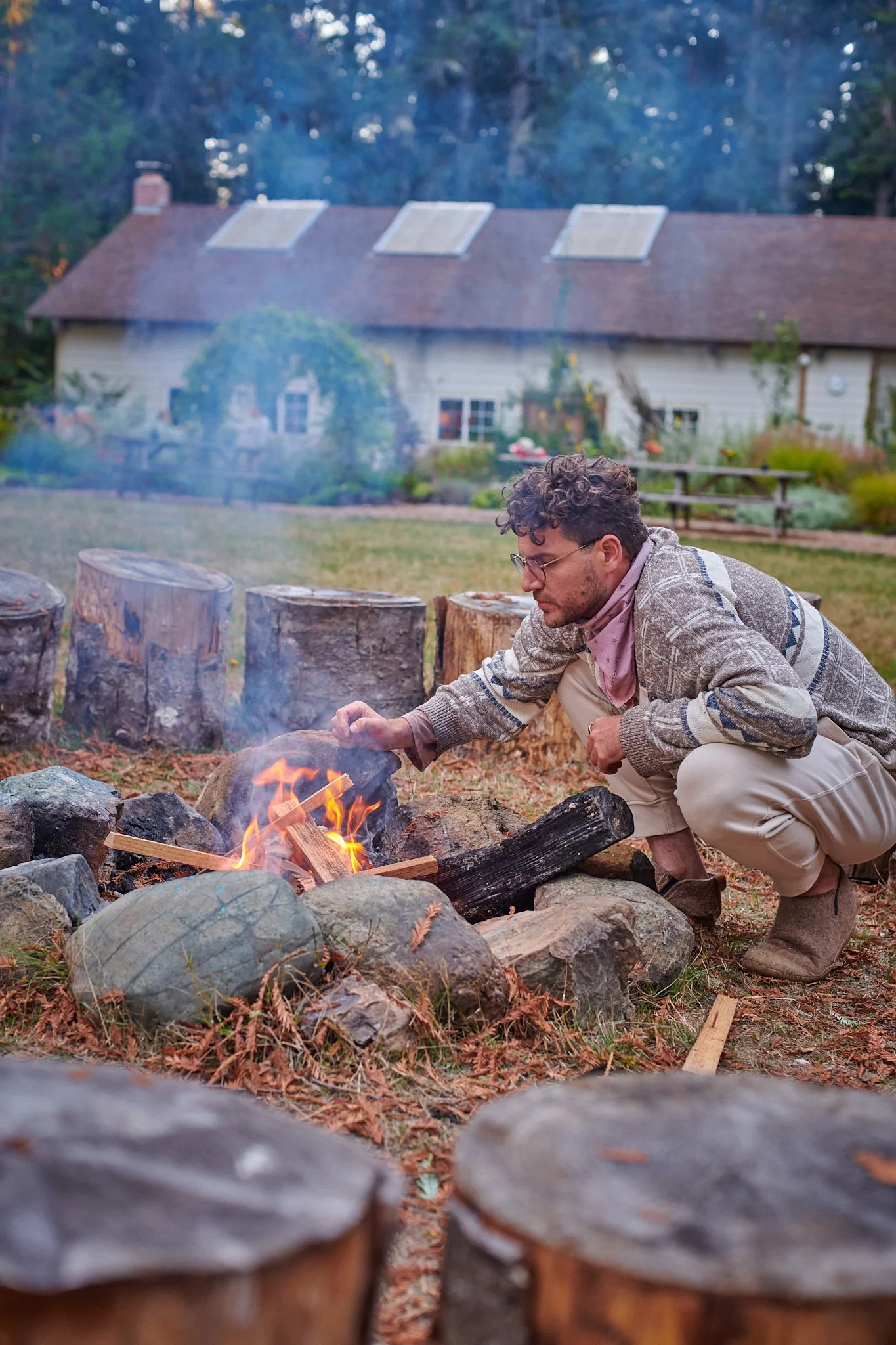 A man crouches near a campfire, tending to the flames with a piece of wood in a backyard with a house and trees in the background.