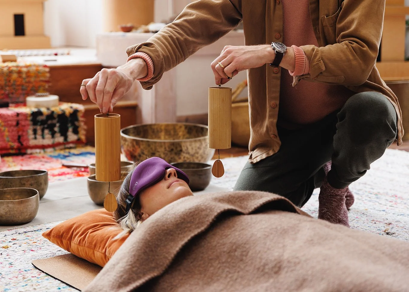 Person lying on a blanket with a sleep mask, receiving sound therapy with hanging bowls from a practitioner in a cozy setting.