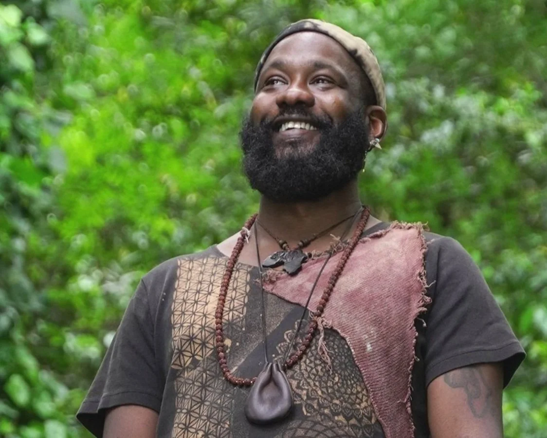 A smiling man with a beard and earrings outdoors, wearing a headwrap and necklaces, with a background of dense green foliage.