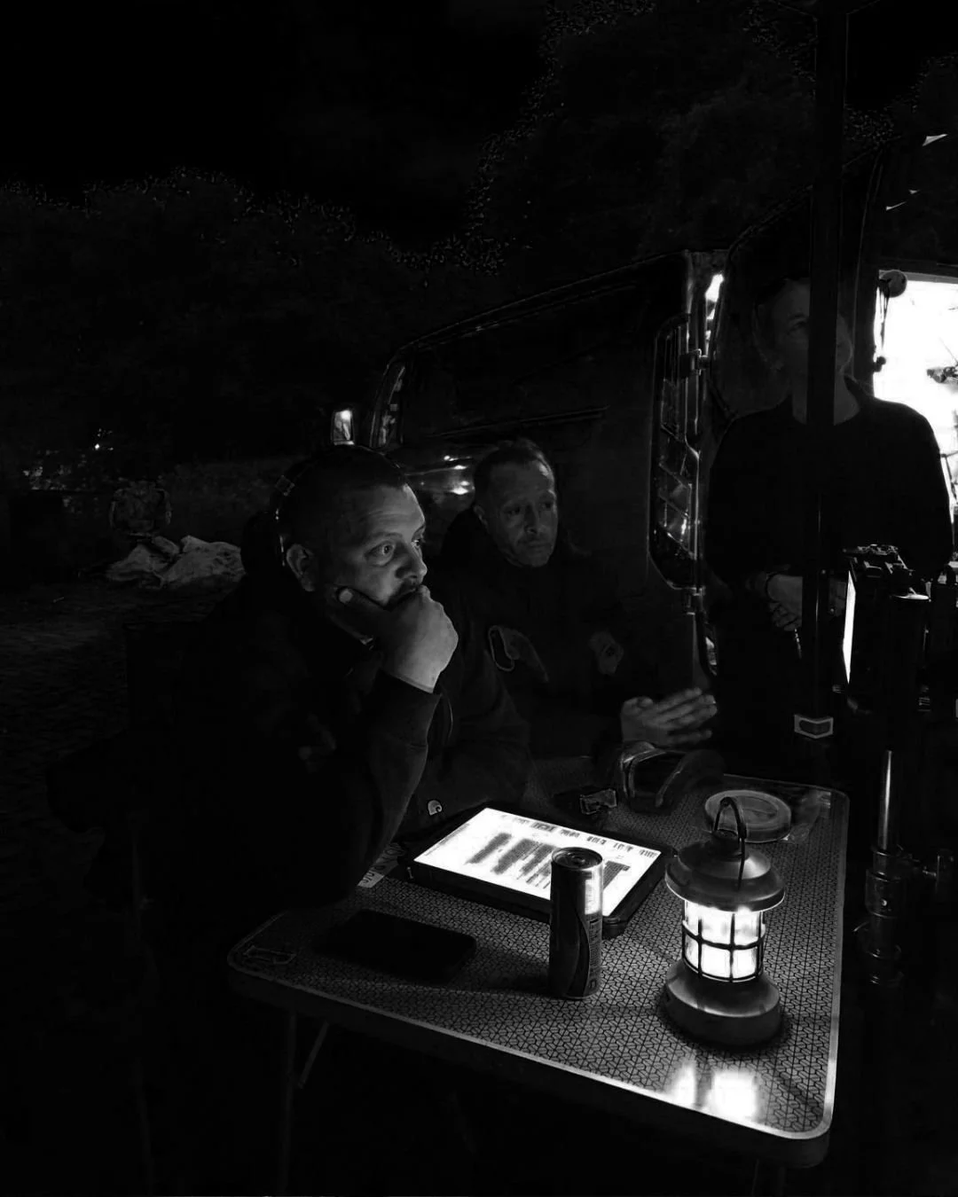 Drie mensen zitten in een donker café of restaurant bij een tafel met een lamp en een digitaal menu, één persoon lijkt te praten terwijl de andere luistert.