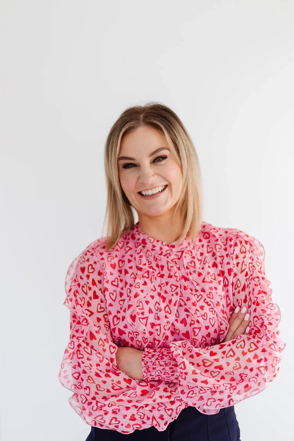 A smiling woman with blonde hair crossing her arms, wearing a pink blouse with red hearts, standing against a plain white background.