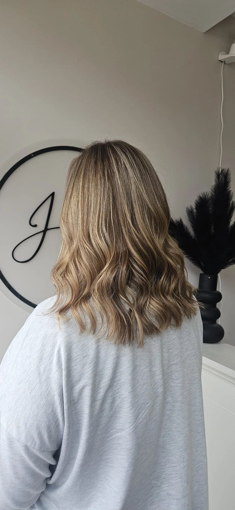 Back view of a woman with shoulder-length, wavy blonde hair, wearing a light gray shirt, standing indoors near a wall with decorative elements including a large black container with black feathers and a sign with the letter 'H'.