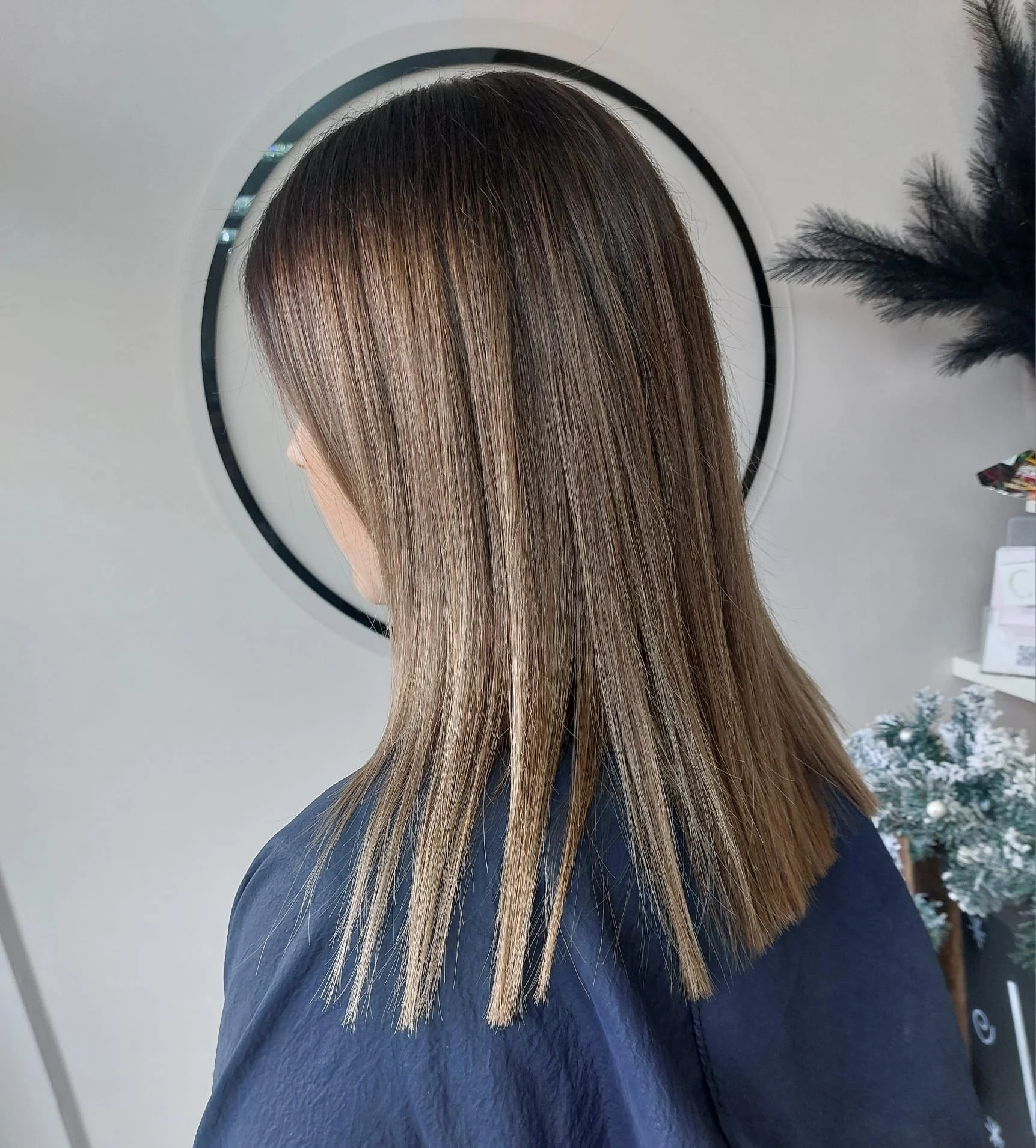 A woman with shoulder-length, straight, light brown hair with darker roots, seen from the back, standing in front of a round mirror in a salon or similar setting.