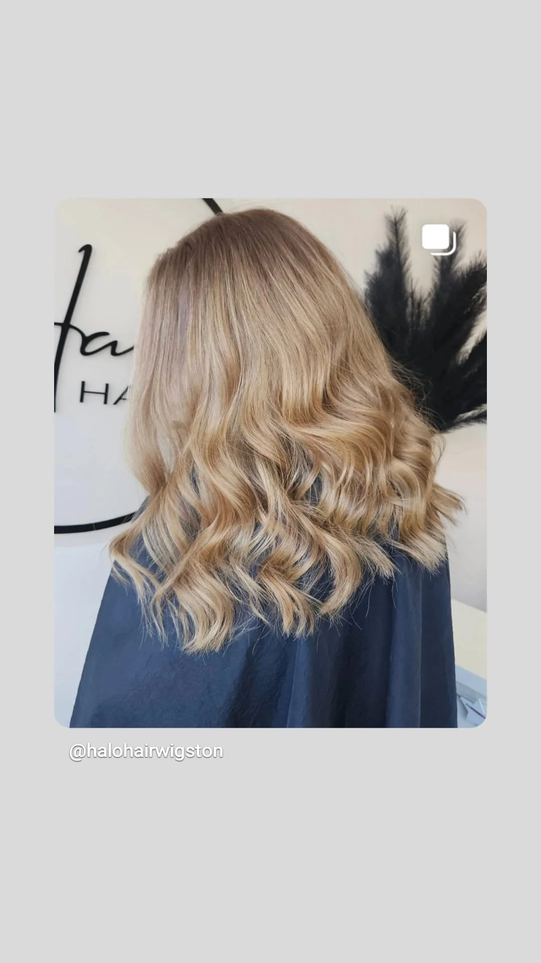 Woman with styled blonde wavy hair in a salon. Black cape around shoulders, salon decor in background.