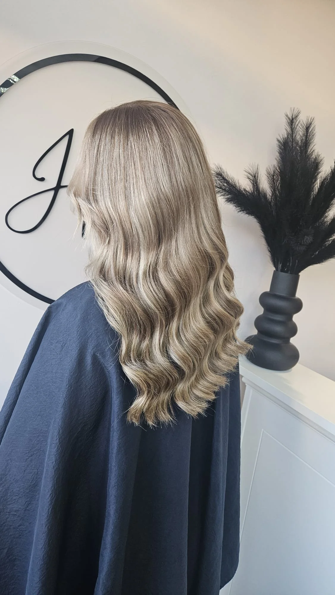A woman with long, wavy blonde hair sitting in a salon, with a black cape over her shoulders, in front of a white wall with a decorative black vase holding black feathers.