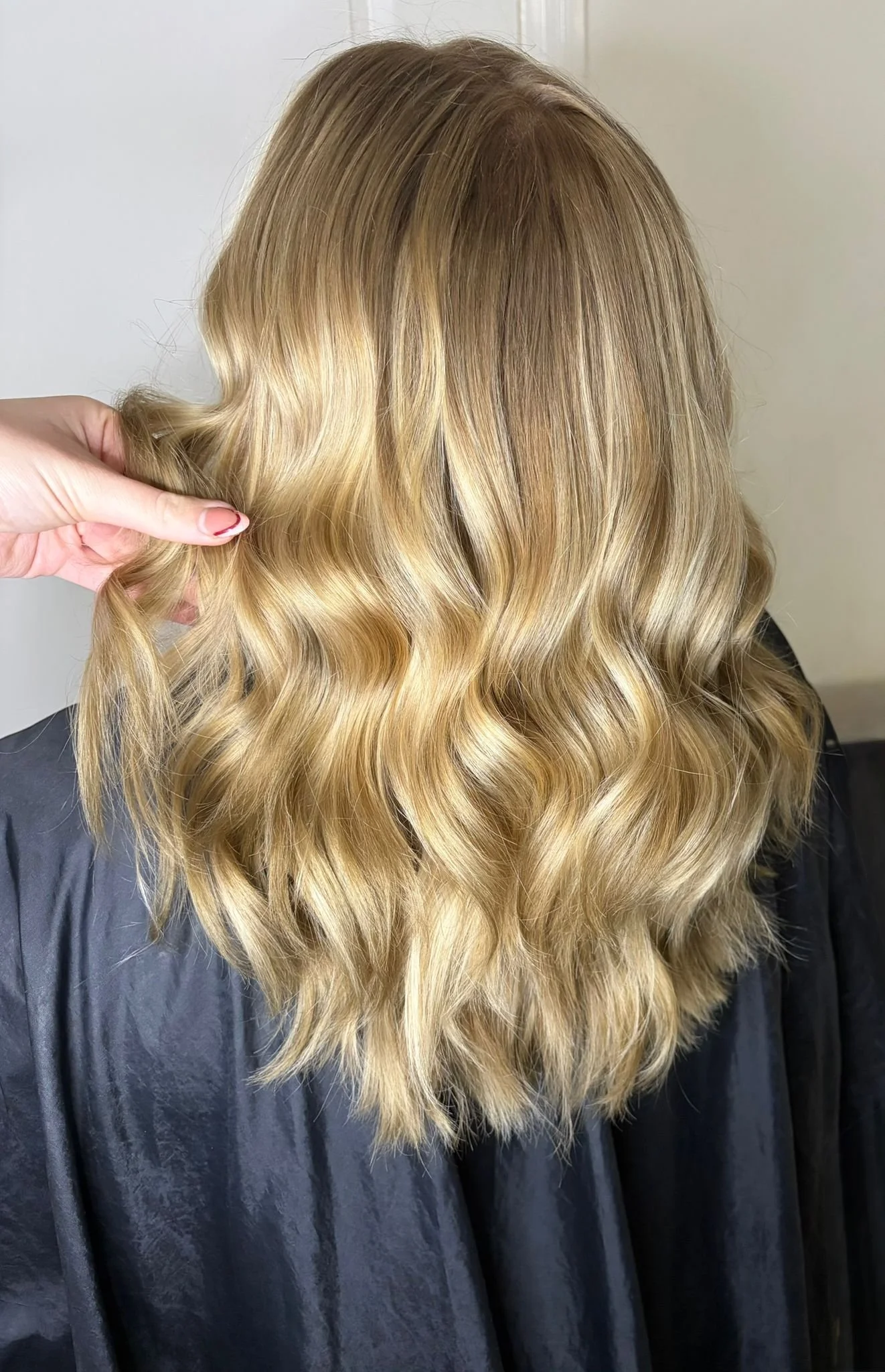 Back view of a woman with wavy blonde hair in a salon, wearing a black cape.