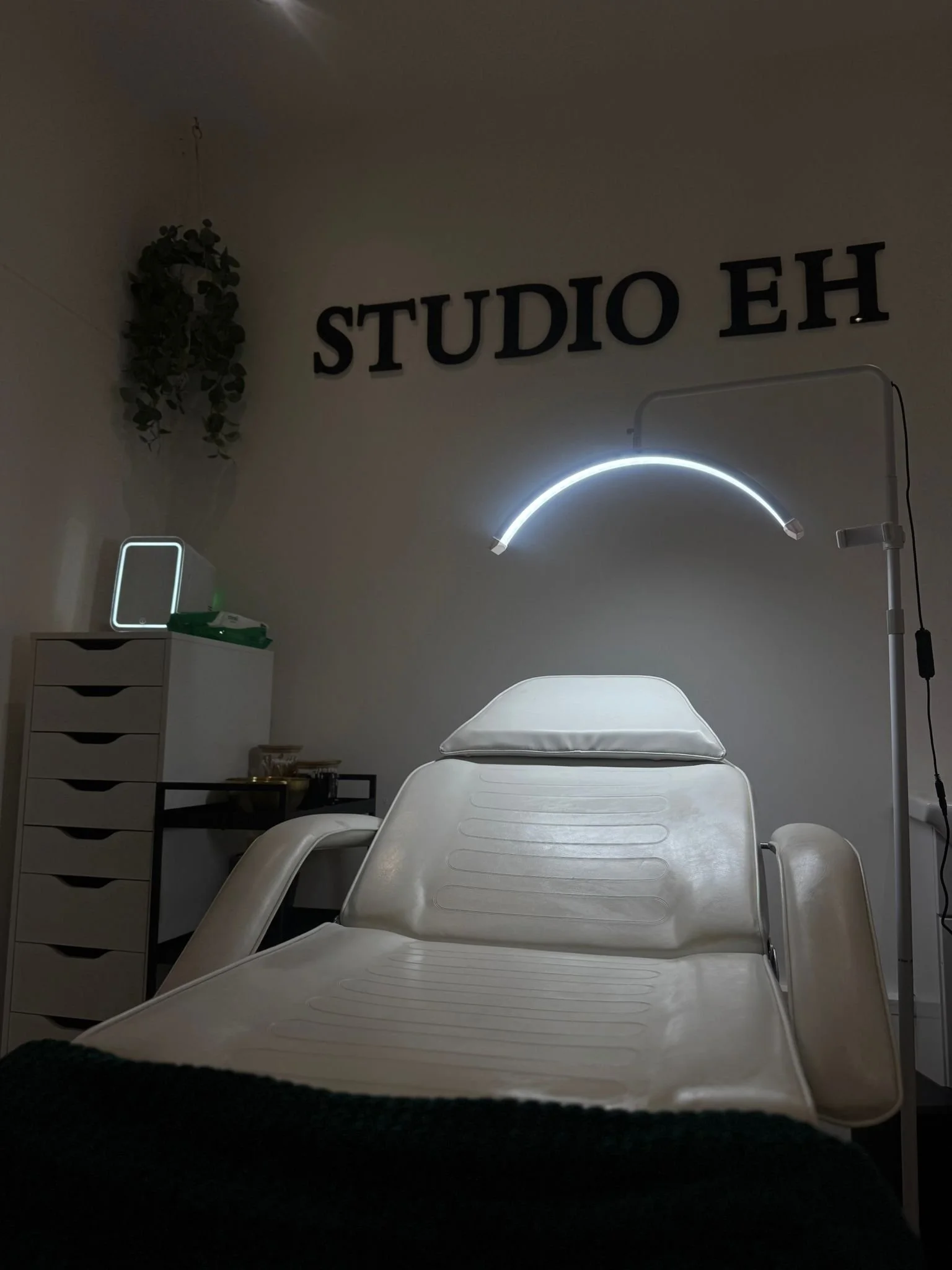A medical treatment or examination room with a white reclining chair, a lamp with a curved light, a sign that reads 'STUDIO EH' on the wall, a cabinet with drawers, and a small mounted mirror or light on top of the cabinet, with some green plants hanging on the wall.