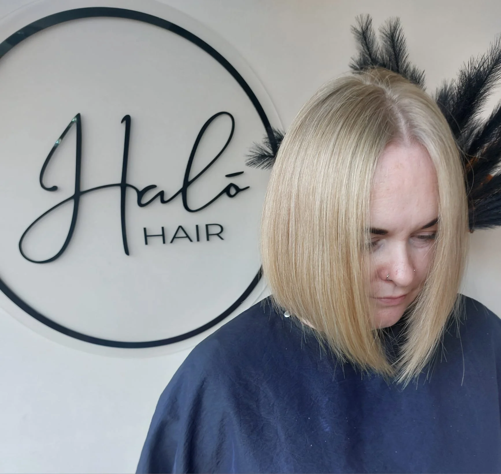 Woman with blonde, straight, shoulder-length hair at a hair salon with a sign that says 'Hala Hair' and black feather decoration in the background.