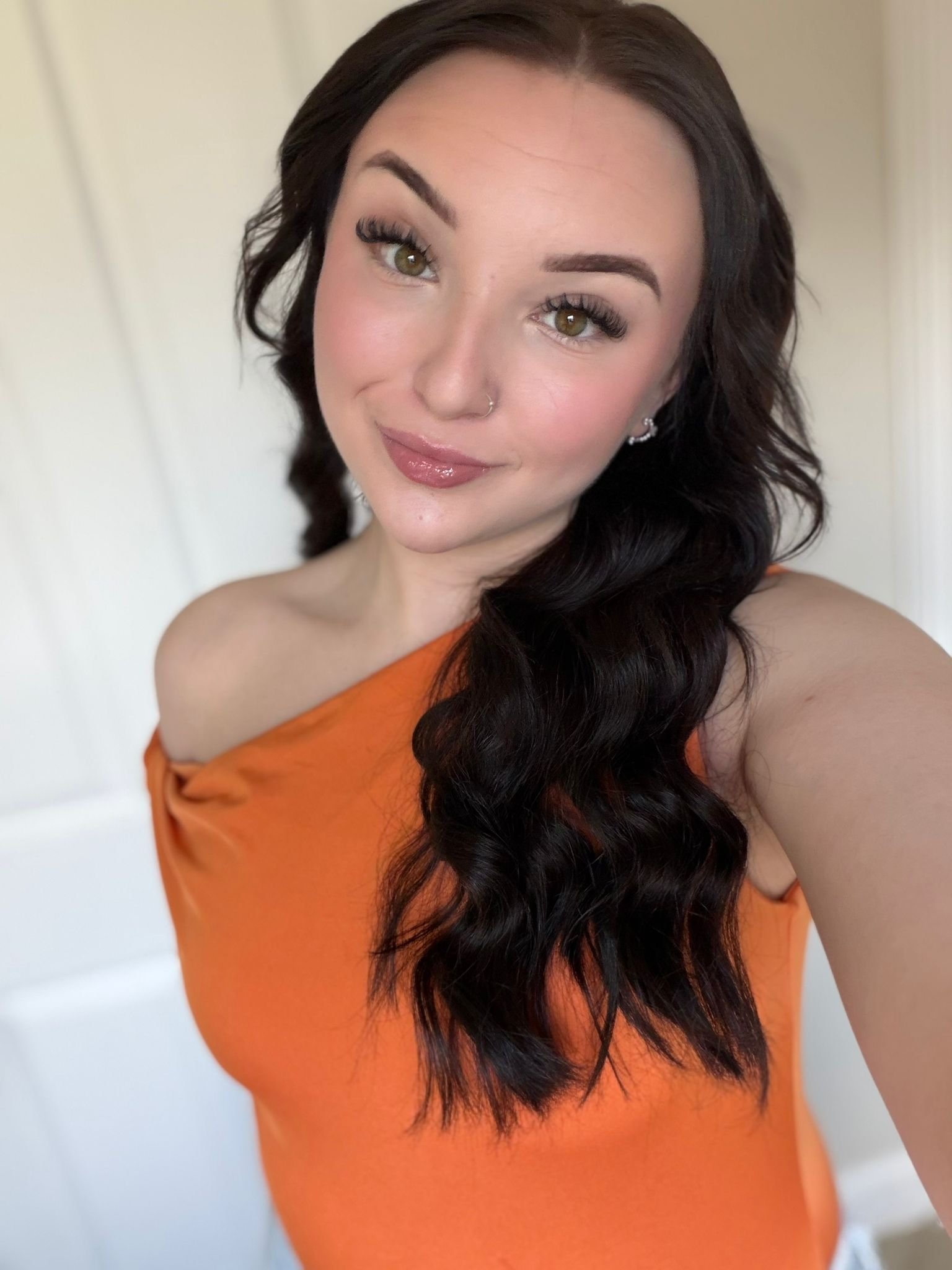 A young woman with wavy dark brown hair, fair skin, and green eyes, smiling with a neutral lipstick and makeup, wearing a one-shoulder orange top, standing indoors with a light-colored background.