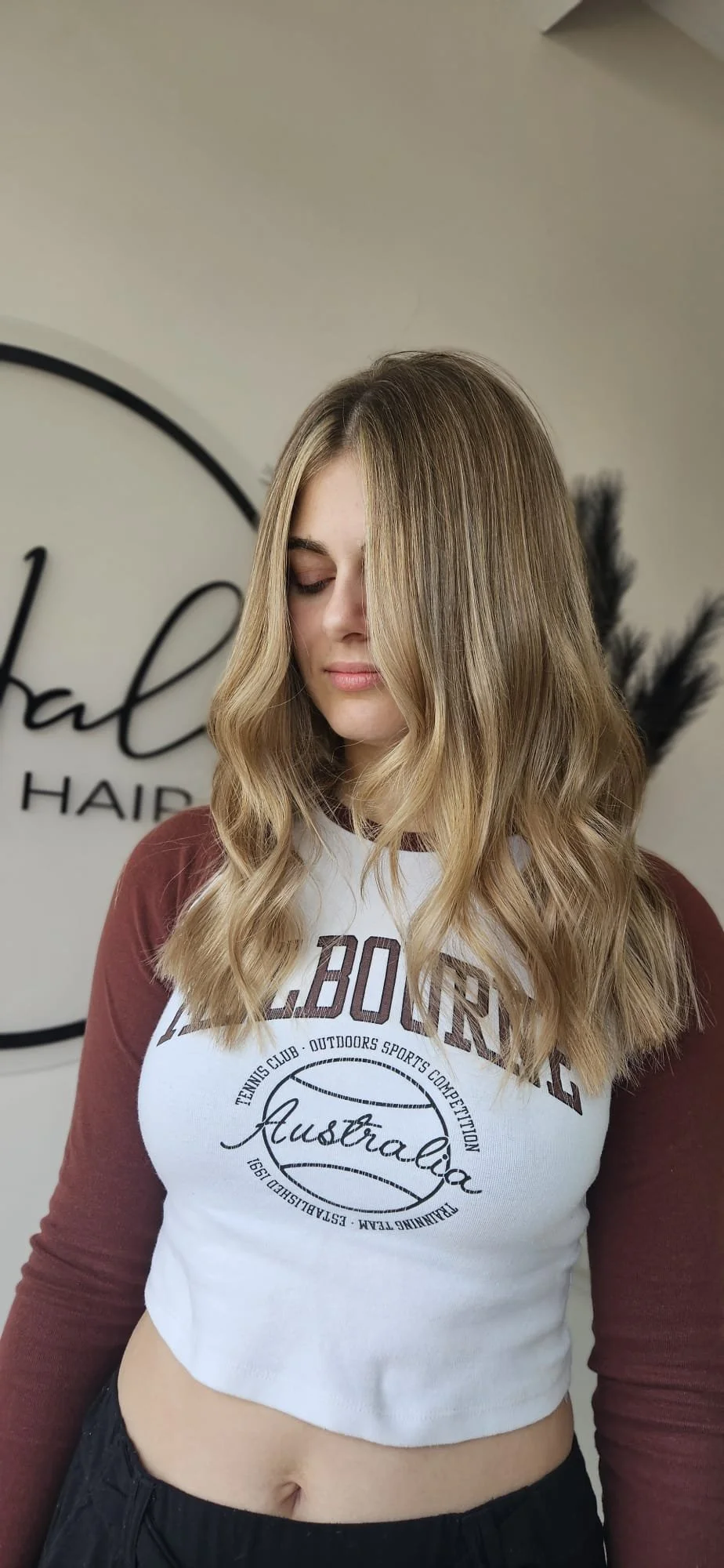 A young woman with wavy blonde hair and light makeup, looking down. She wears a white and maroon long-sleeve crop top with the words 'L.B.ORY C' and 'Australia' printed on it, and black pants. A salon logo is visible in the background.