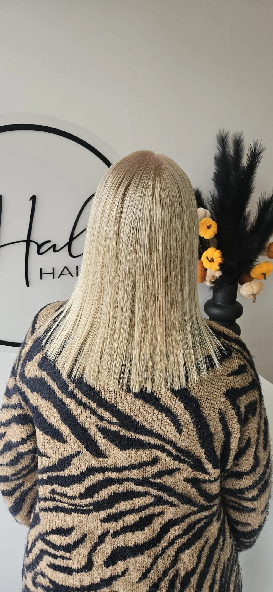 Back view of a woman with straight blonde hair wearing a beige and black zebra-patterned sweater, standing in front of a wall with a black and white logo and a flower arrangement with yellow and black flowers.