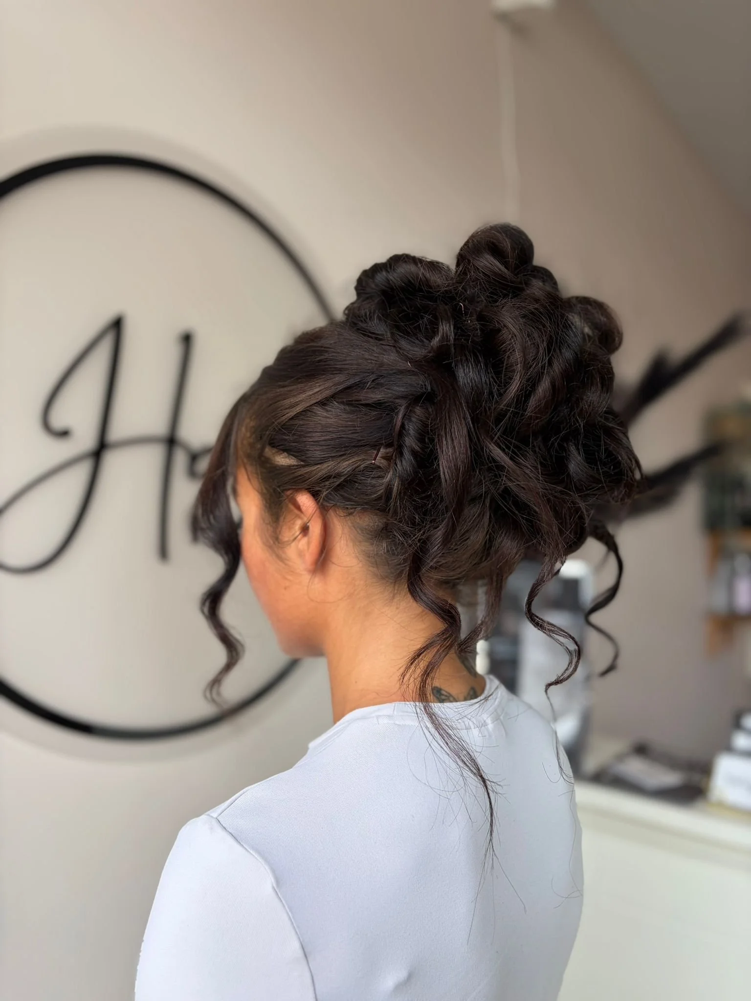 A woman with dark brown hair styled in an elaborate updo with curls, wearing a white top, in a salon with a logo on the wall