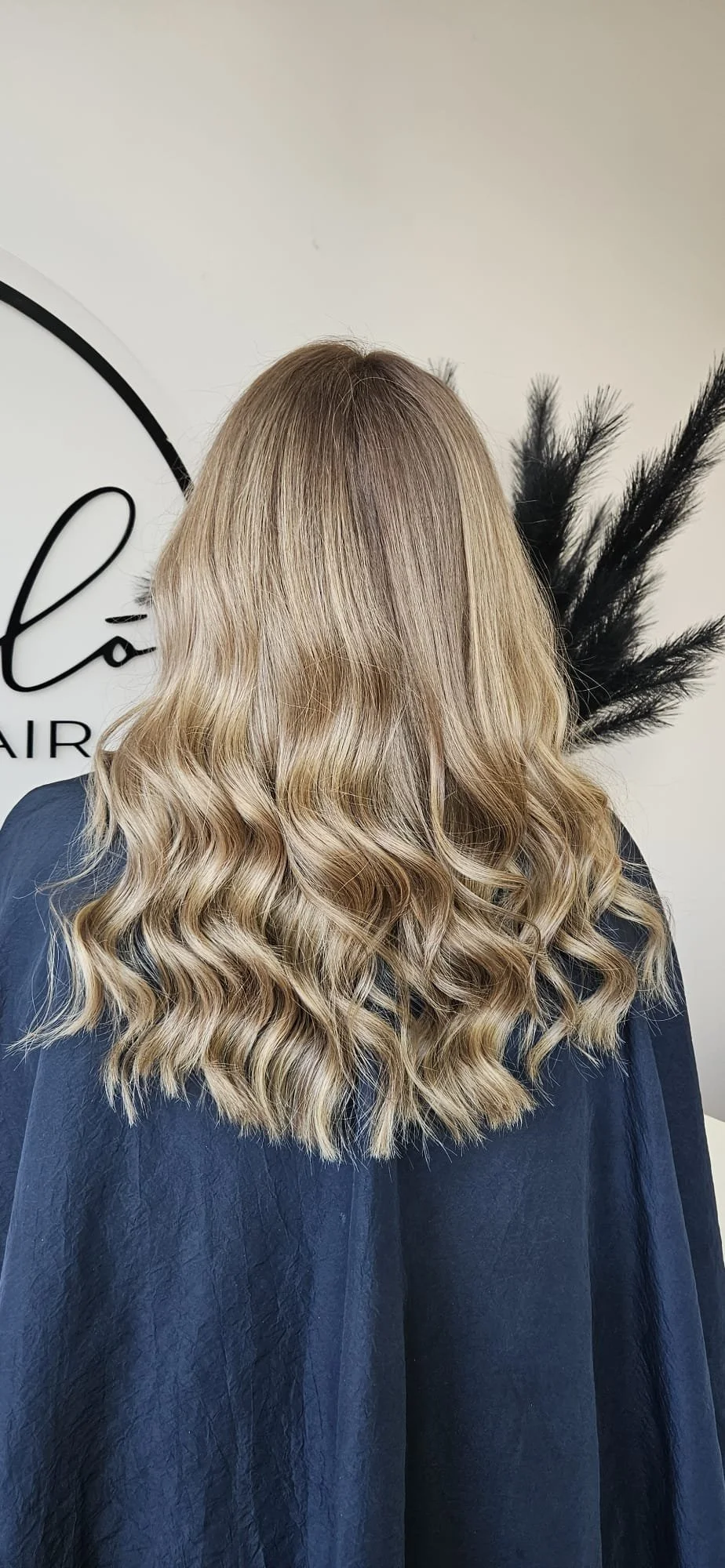 Back view of a woman with long, wavy blonde hair in a salon setting.