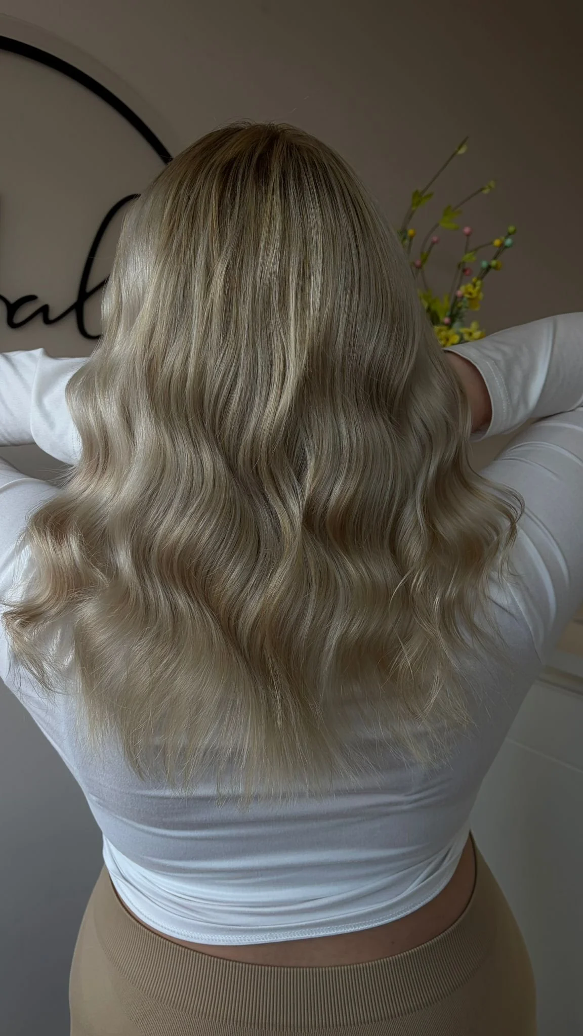 Back view of a woman with long, wavy blonde hair wearing a white top and beige pants.