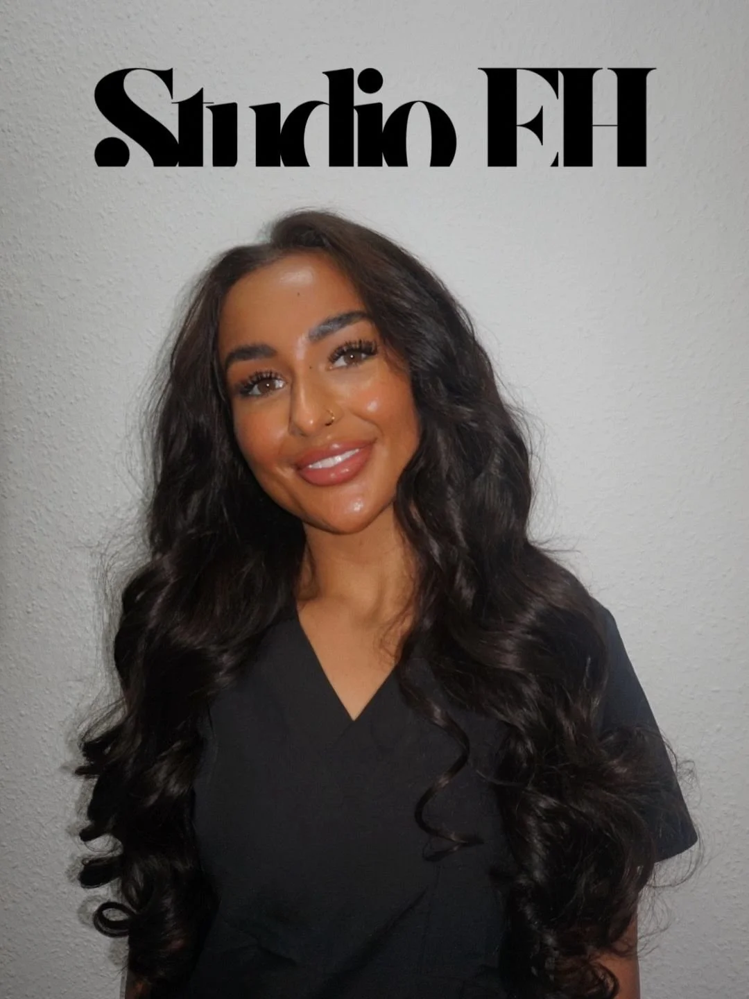 A woman with long, dark, wavy hair, smiling, wearing a black top, standing against a white wall with the words 'Studio EH' written upside down above her.
