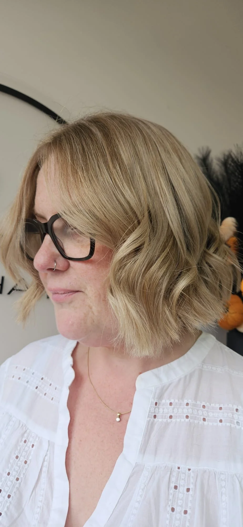 A woman with blonde, wavy, shoulder-length hair and glasses, wearing a white top with eyelet details and a delicate necklace, in a close-up side profile shot.