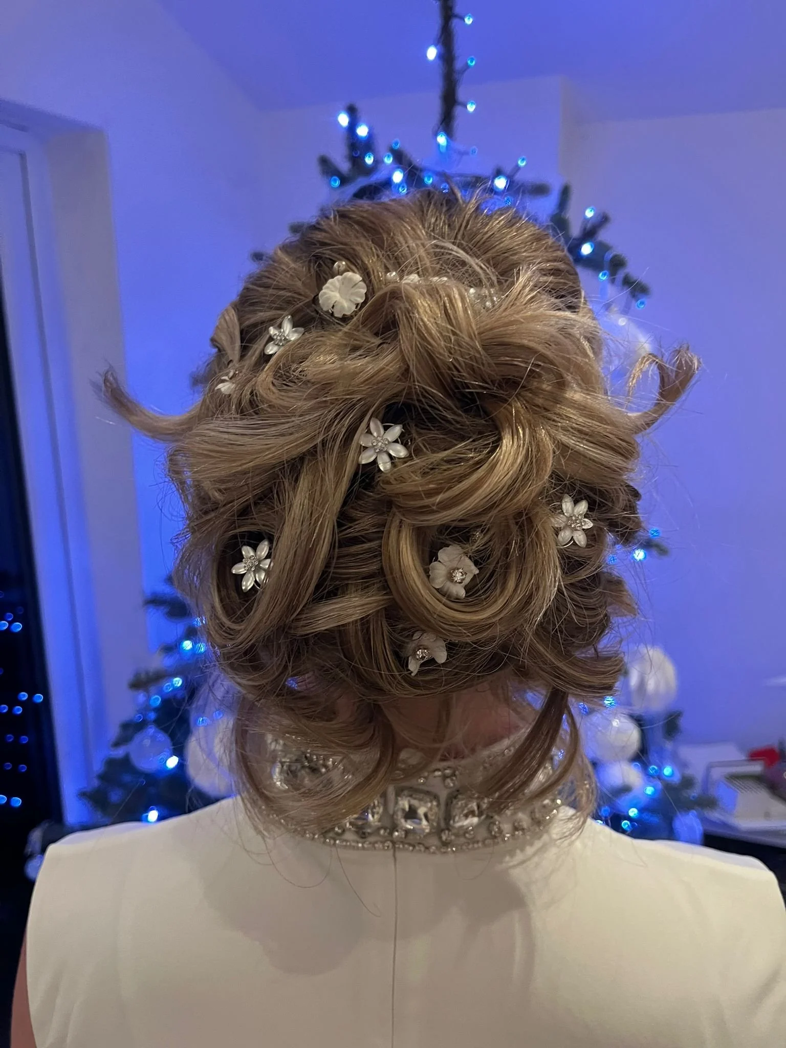 Updo bridal hairstyle decorated with white floral hairpins, with a decorated neckline visible, in front of a Christmas tree with blue and white lights.