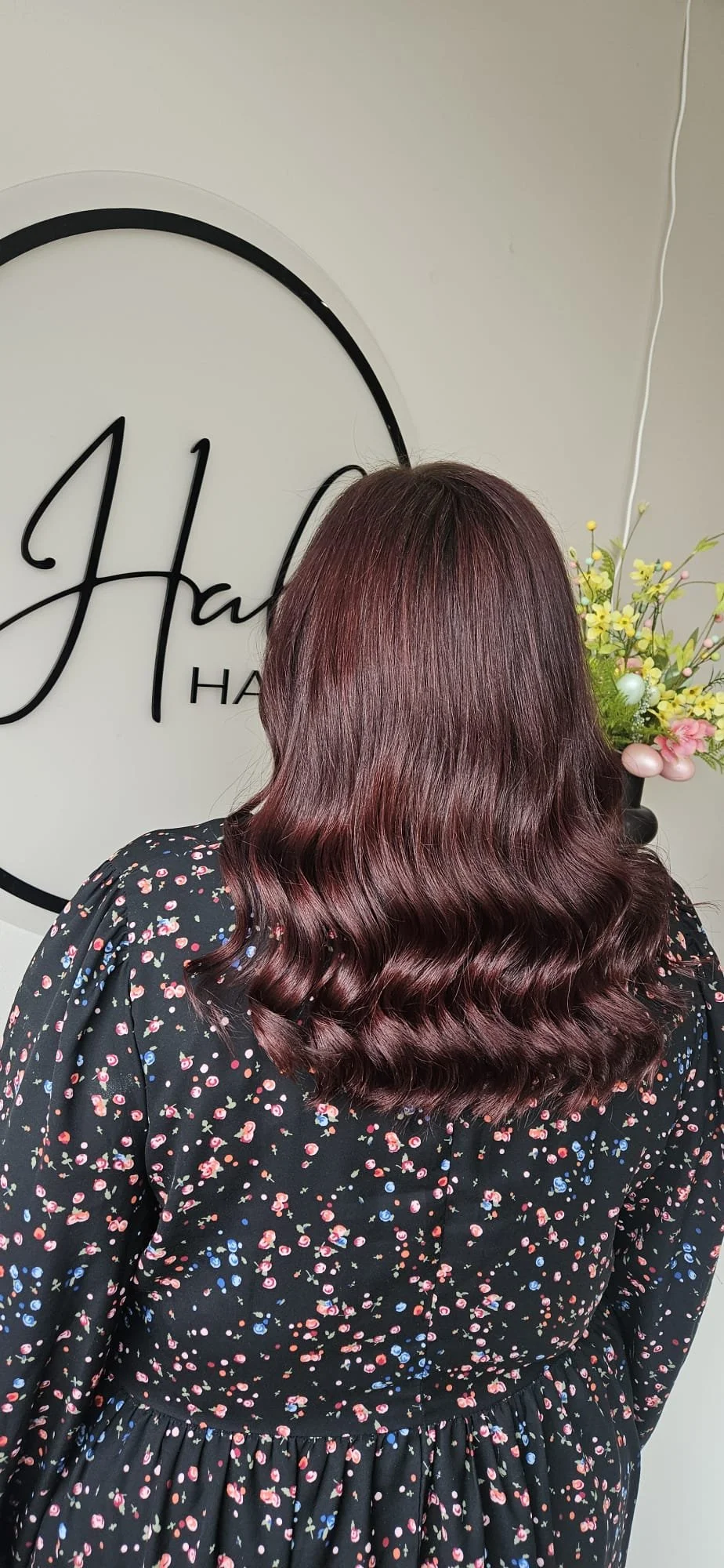 Back view of a woman with medium-length, wavy, burgundy hair standing in front of a wall with the word 'Halo' written on it and a vase with flowers.
