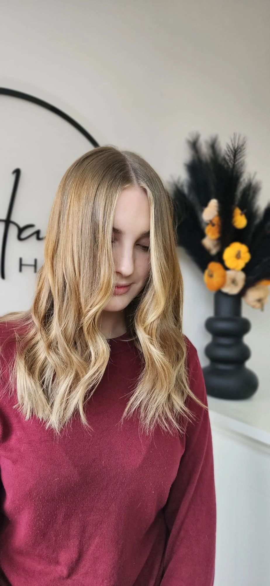 A young woman with wavy blonde hair and a maroon sweater, eyes closed, standing indoors with a decorative black vase and dried flower arrangement in the background.
