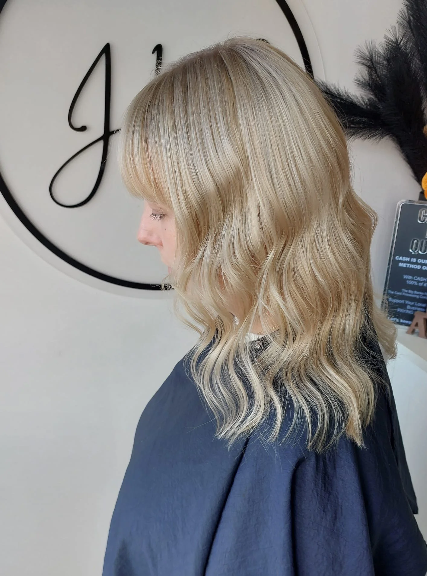 Side profile of a woman with blonde wavy hair wearing a black salon cape, standing in front of a wall with a large decorative clock.