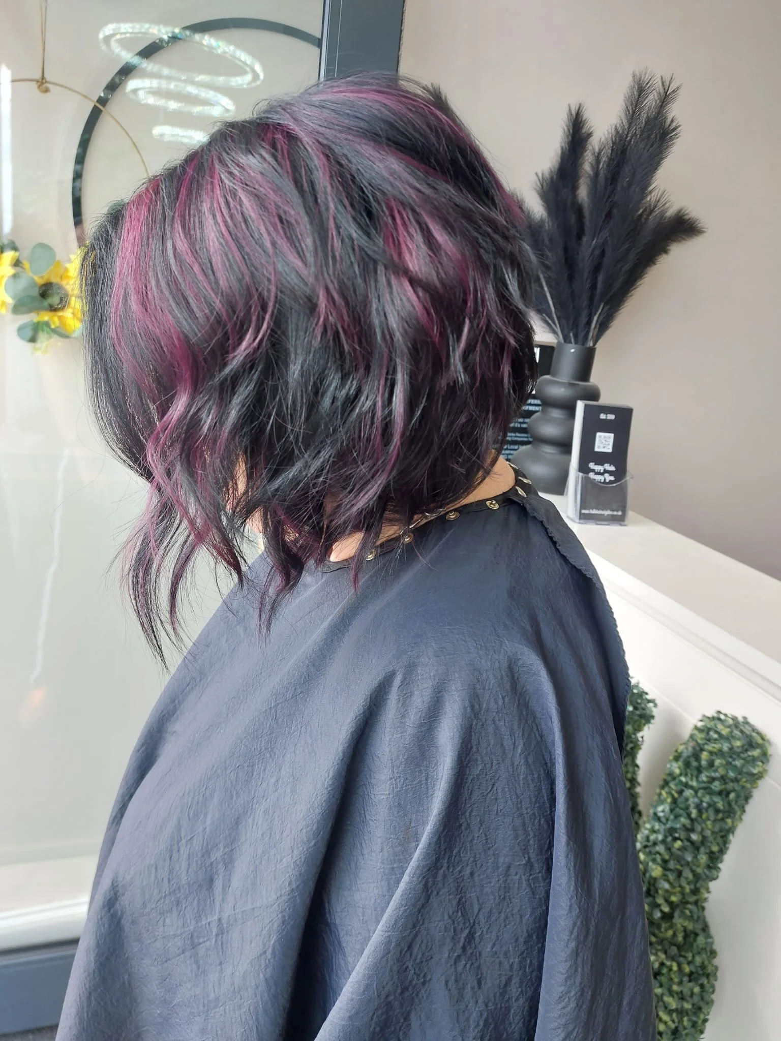 A person with shoulder-length wavy black hair with pink highlights, wearing a black cape, at a salon or hair studio.