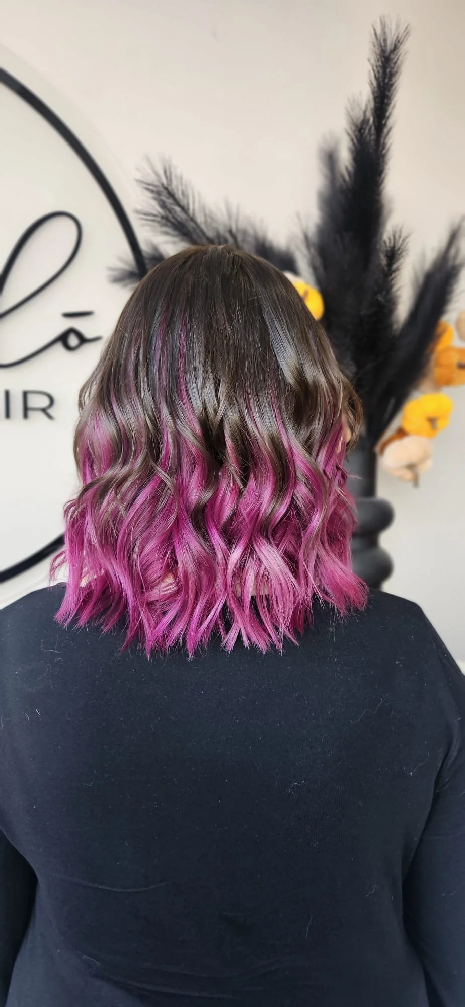 Back of woman with wavy hair that is dark at the roots and pink at the ends, wearing a black shirt, standing in front of a wall with decorative black feathers and a large circular logo.
