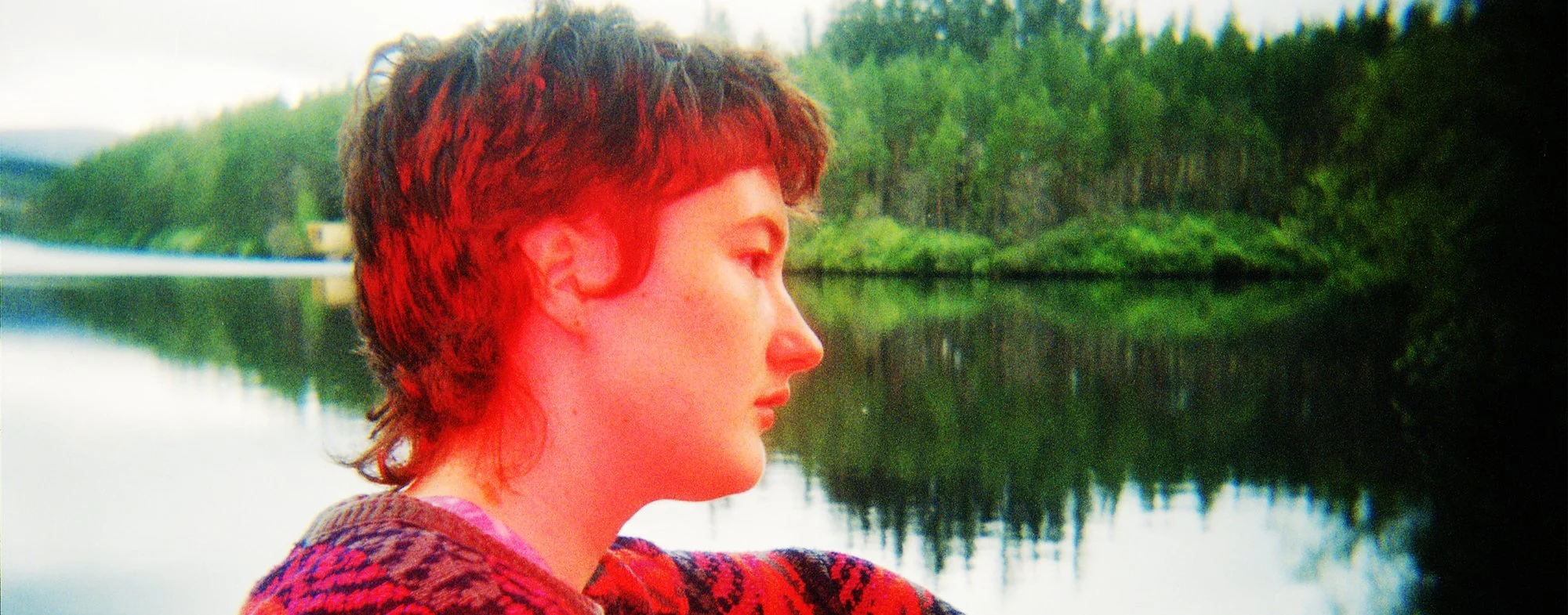 A woman with short, curly red hair looking to the right near a body of water surrounded by green trees.
