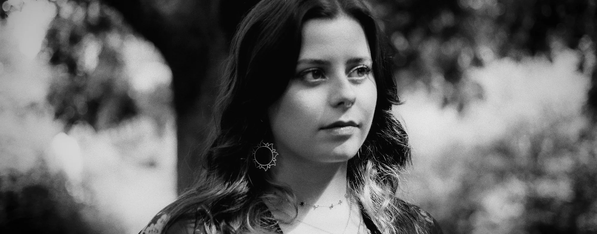 A woman with dark hair and light skin, wearing earrings, looking contemplative outdoors.