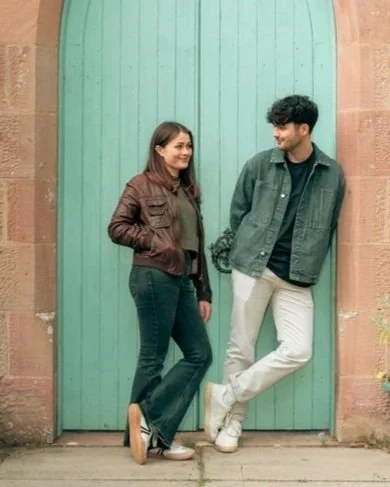 A young woman and young man standing in front of a large turquoise door, smiling and looking at each other.