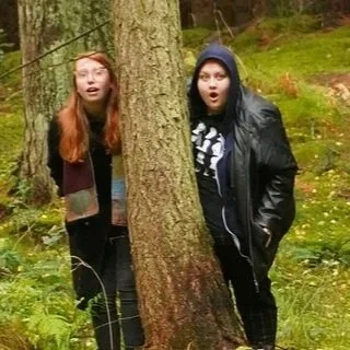 Two women peeking from behind a tree in a forest.