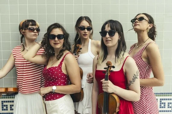 Five women with dark sunglasses posing against a tiled wall, some holding violins.