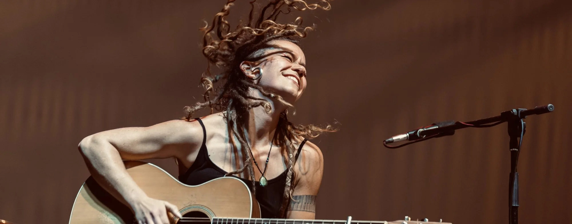 A woman with long dreadlocks playing an acoustic guitar and smiling on stage, with a microphone in front of her.