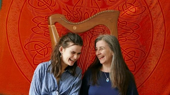 Two women smiling and laughing together in front of a red decorative background with Celtic knot patterns, one wearing a blue shirt and the other wearing a dark blue top.