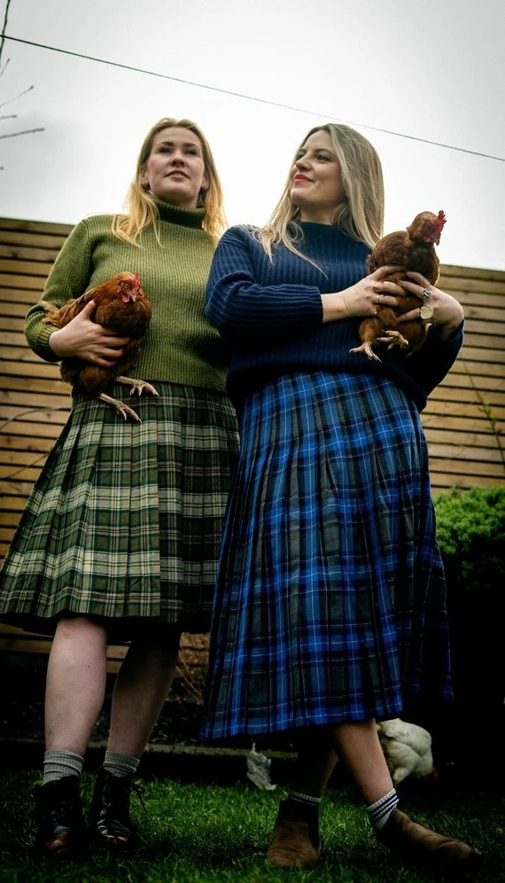Two women standing outdoors, each holding a chicken, wearing plaid skirts and sweaters, with a wooden fence and cloudy sky in the background.