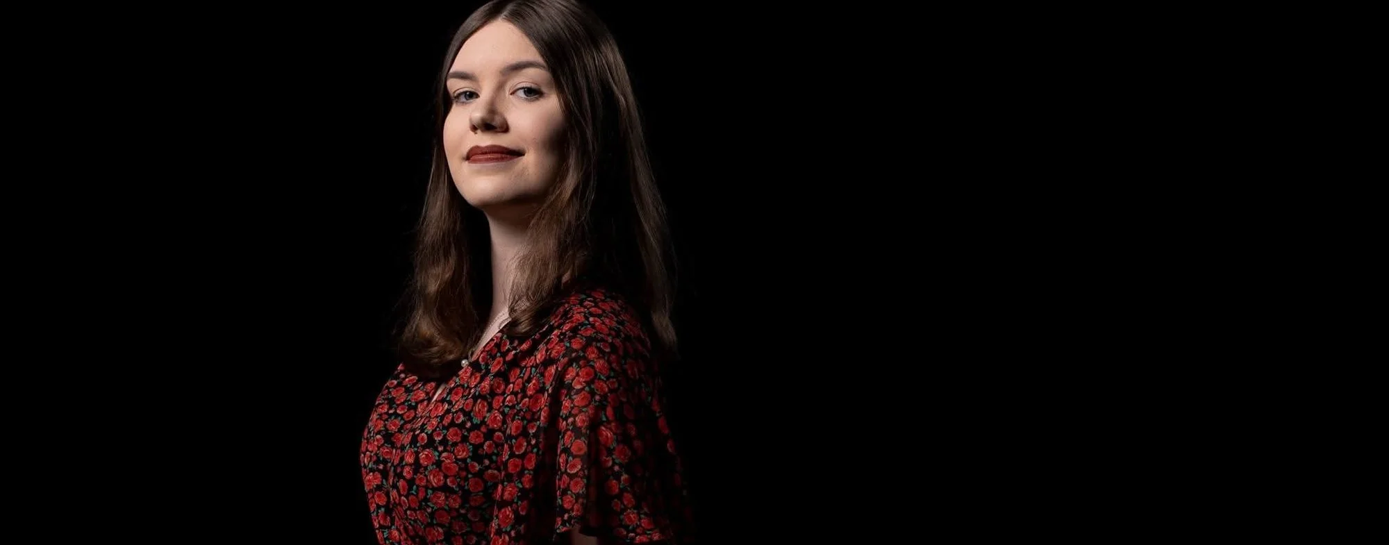 A young woman with straight brown hair, wearing a red and black patterned dress, posing against a black background.