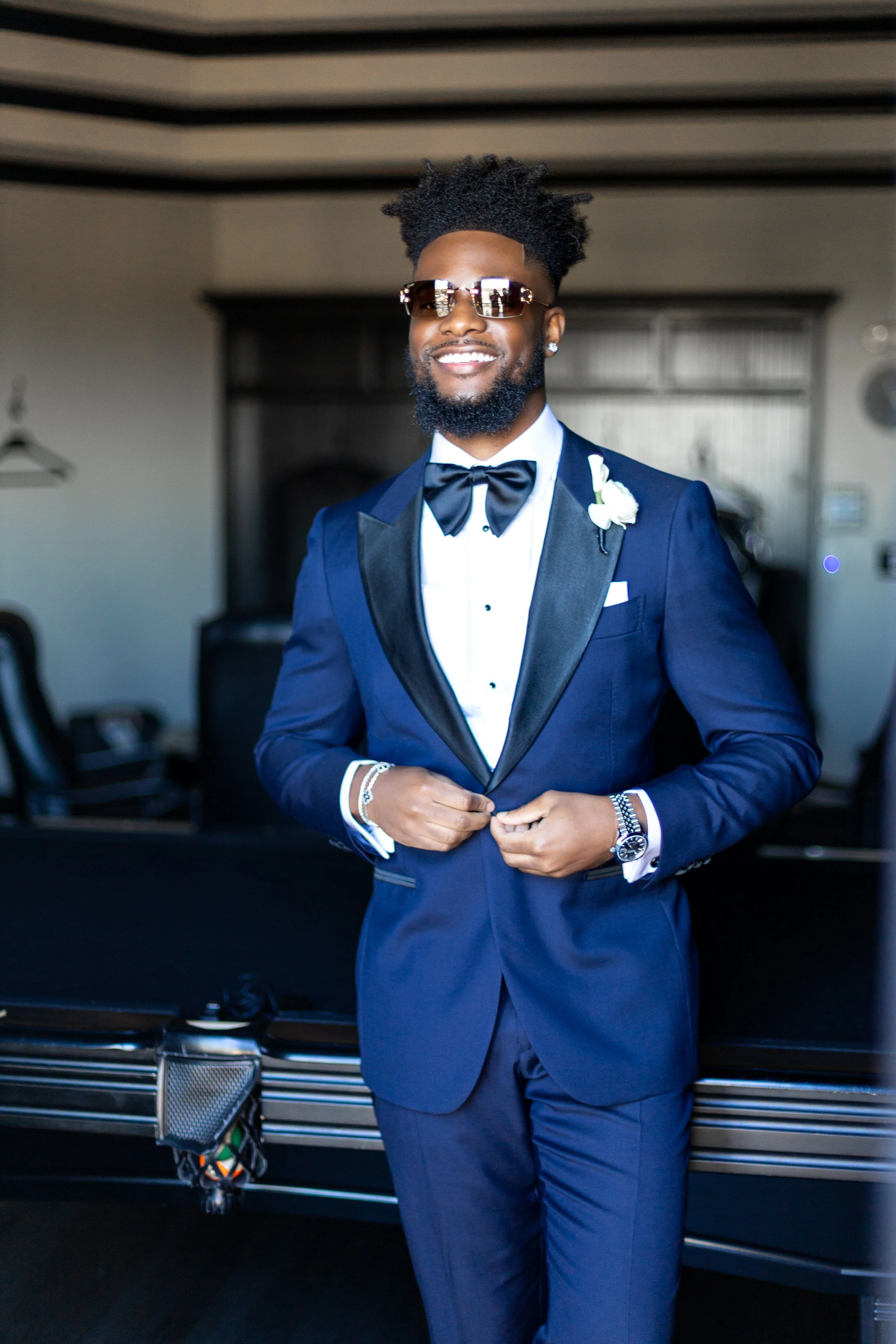 A man in a blue tuxedo with black lapels, white shirt, black bow tie, sunglasses, and a wristwatch, smiling and adjusting his jacket.