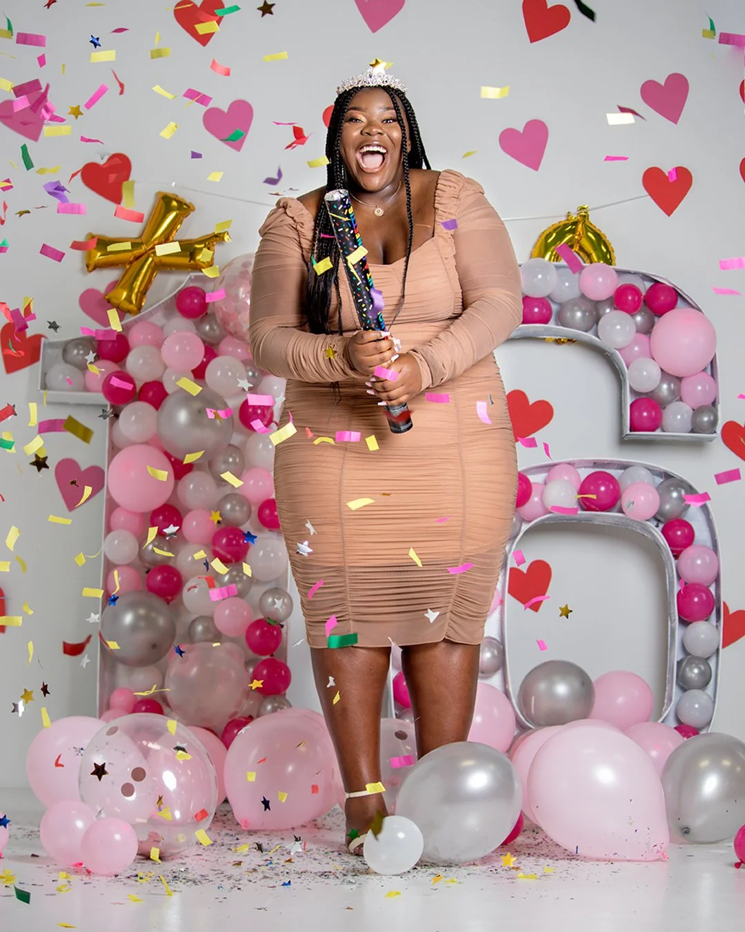 A woman wearing a beige dress celebrating with balloons, confetti, and decorations, holding a party popper, surrounded by pink, white, gray, and gold balloons, with a large number 6 in the background and hearts on the wall.