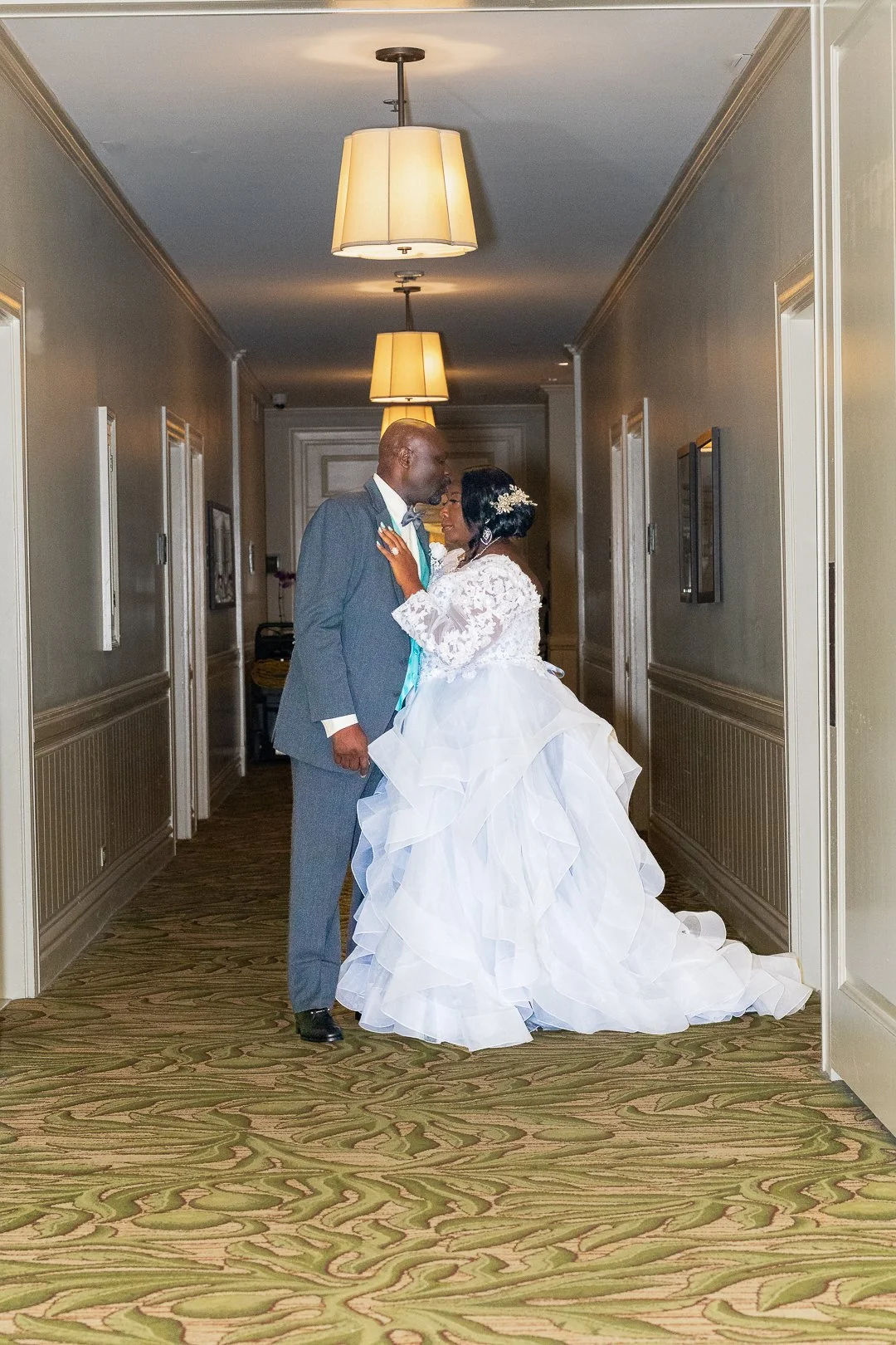 A bride and groom standing in a hotel hallway, touching foreheads and smiling at each other, with warm lighting overhead.