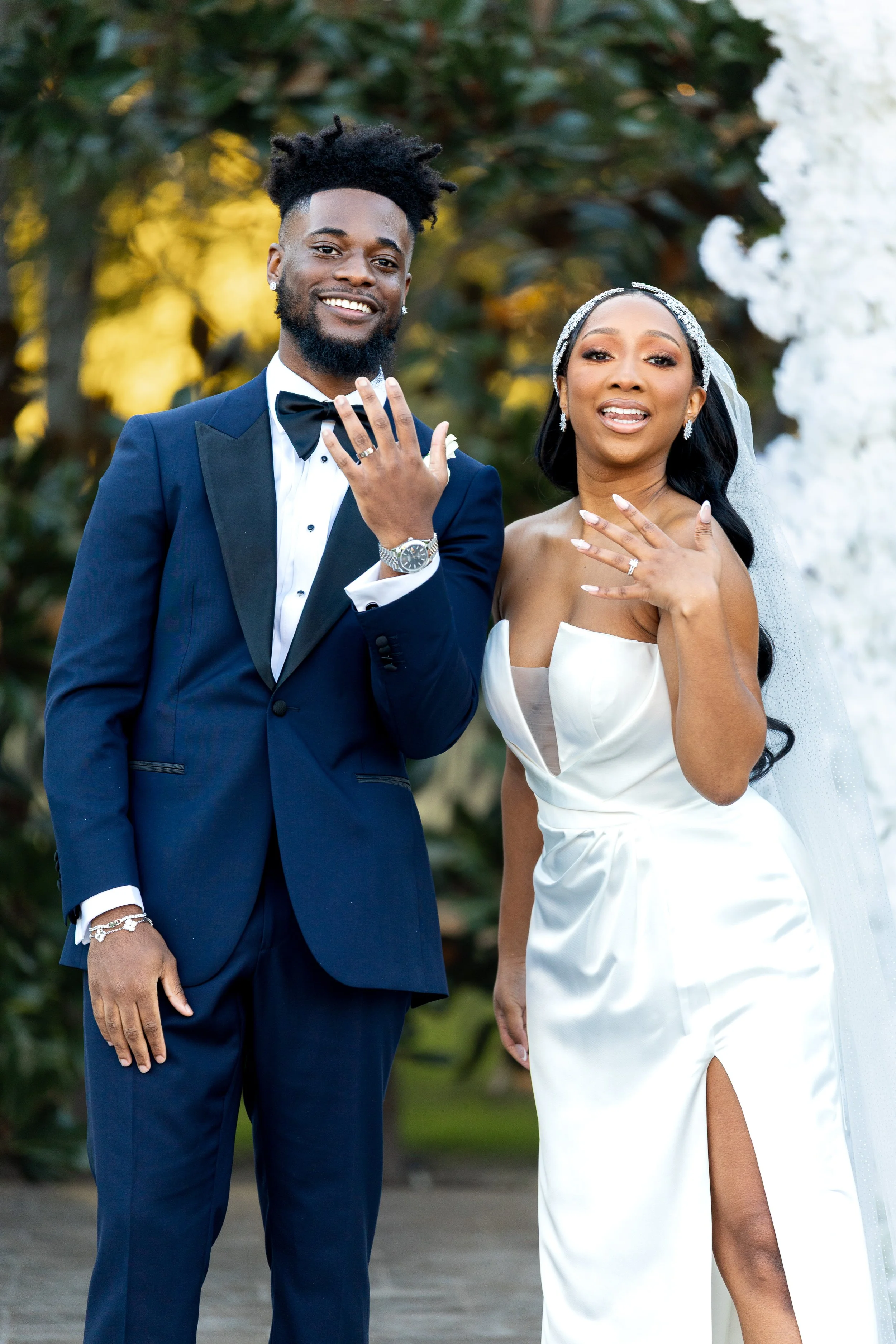 A newlywed couple, dressed in wedding attire, smiles and shows their wedding rings in an outdoor setting with greenery and flowers in the background.