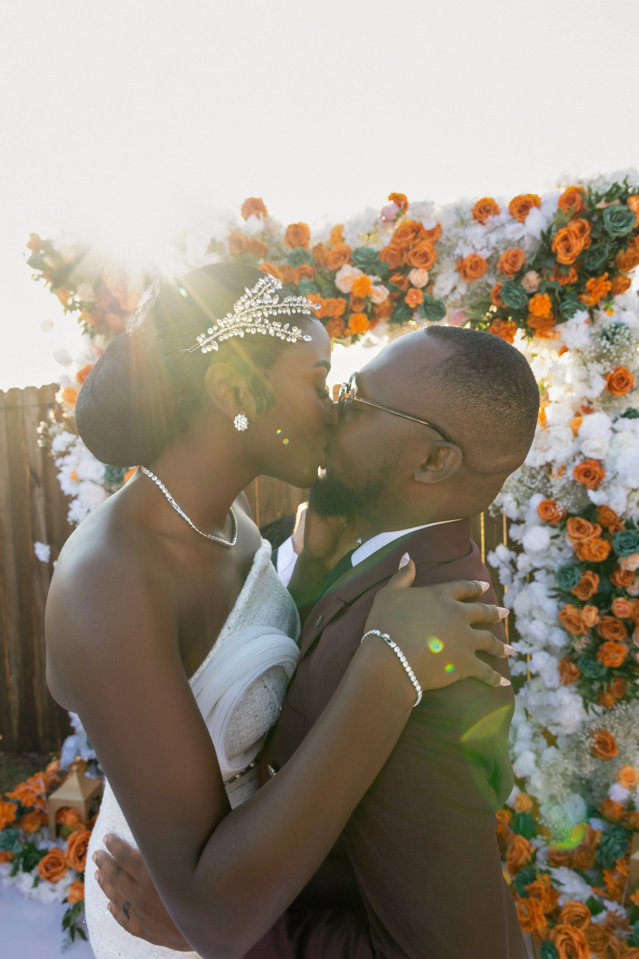 A bride and groom kiss at their wedding, standing in front of a floral arch with orange and white flowers, with sunlight shining behind them.